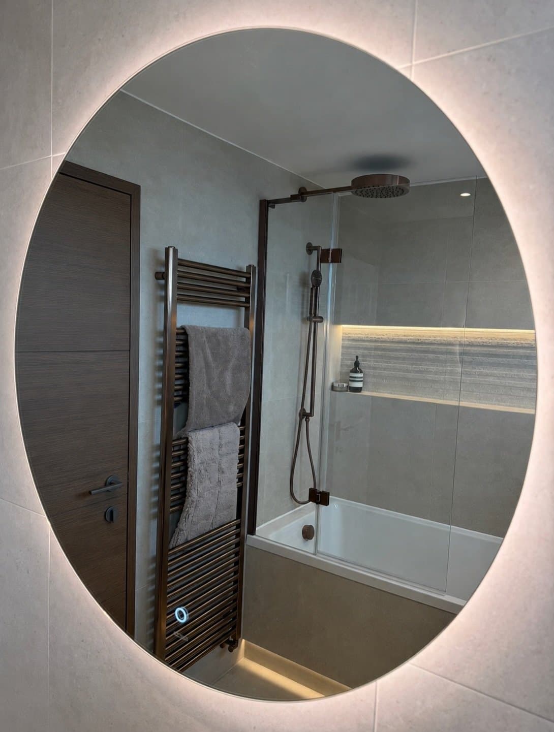 Backlit round mirror reflecting a bronze rain shower and lit stone niche